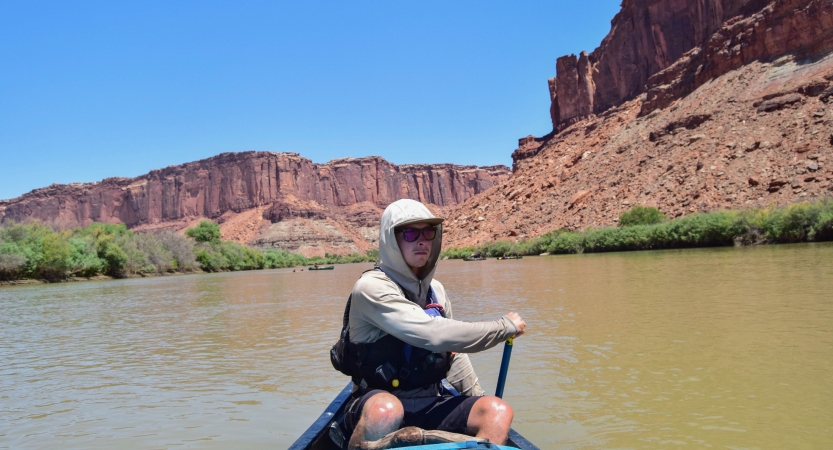 a person sitting at the back of a canoe paddles the watercraft forward. There are tall canyon walls behind them.